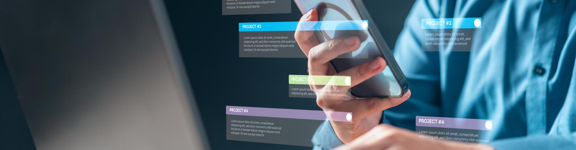 A person in a blue shirt sits at a desk, holding a smartphone in one hand while typing on a laptop with the other; colorful translucent digital project-label overlays float around the phone and laptop screen, suggesting task management or multitasking.
