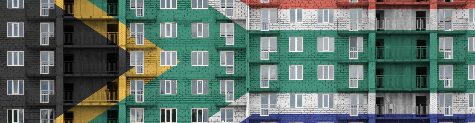 A multi-story apartment building with the South African flag superimposed over it. The building's facade is covered in windows and balconies, and the flag's colors (black, yellow, green, white, red, and blue) are clearly visible.