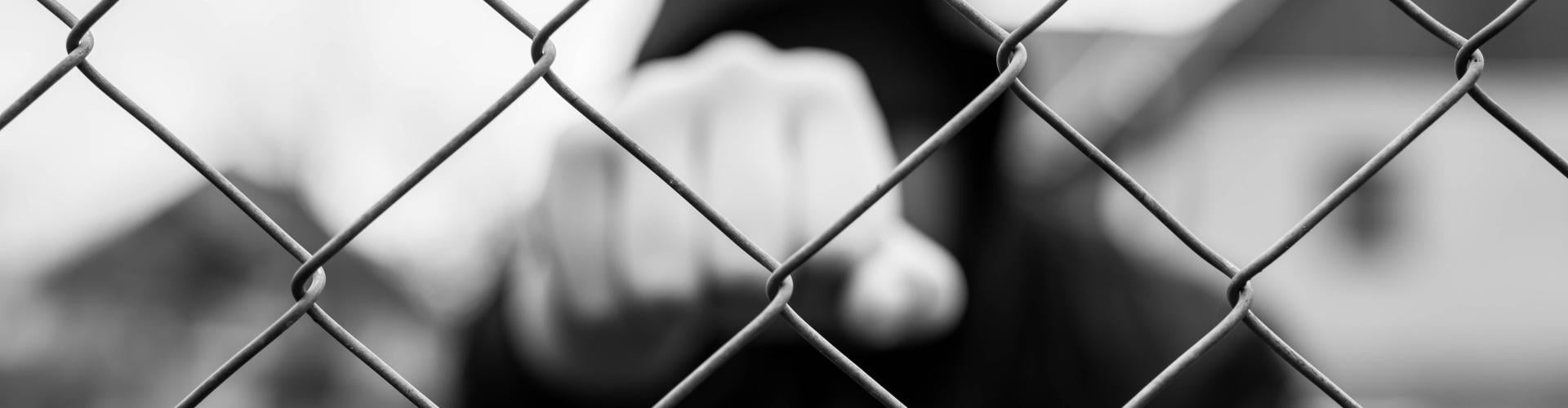 Black-and-white photo of a person wearing a hooded jacket standing behind a chain-link fence; the fence is in sharp focus while the person is blurred and extends a finger toward the camera.