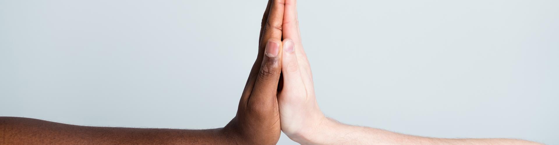 Close-up photo of two outstretched arms, one with dark skin and one with light skin, palms touching in a high-five against a pale gray background.