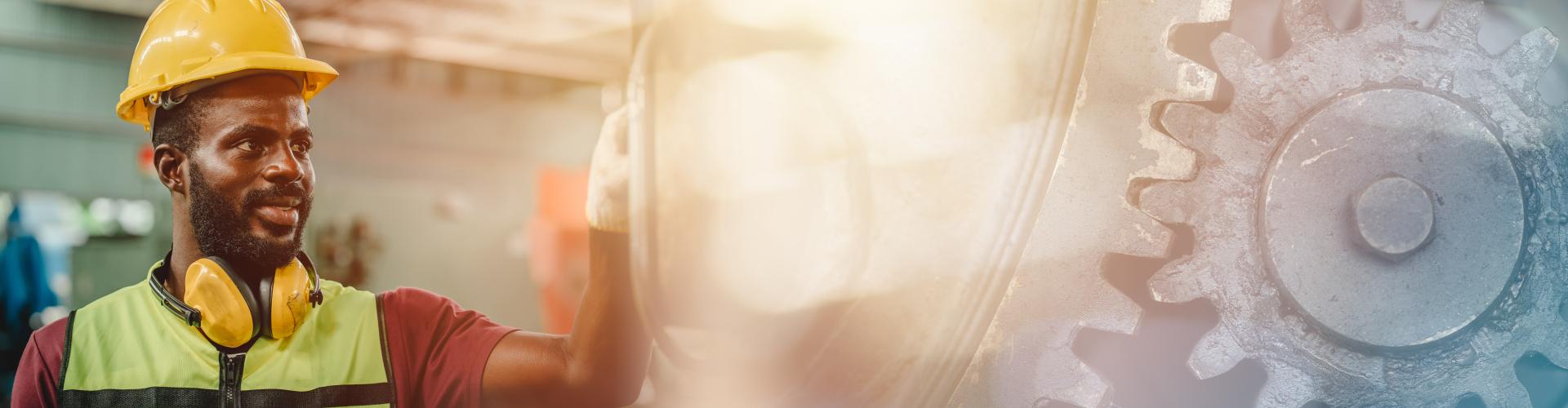 A male factory worker wearing a yellow hard hat, yellow hearing protection around his neck, and a reflective safety vest reaches toward large metal gears inside an industrial workshop; warm sunlight flares in the background.