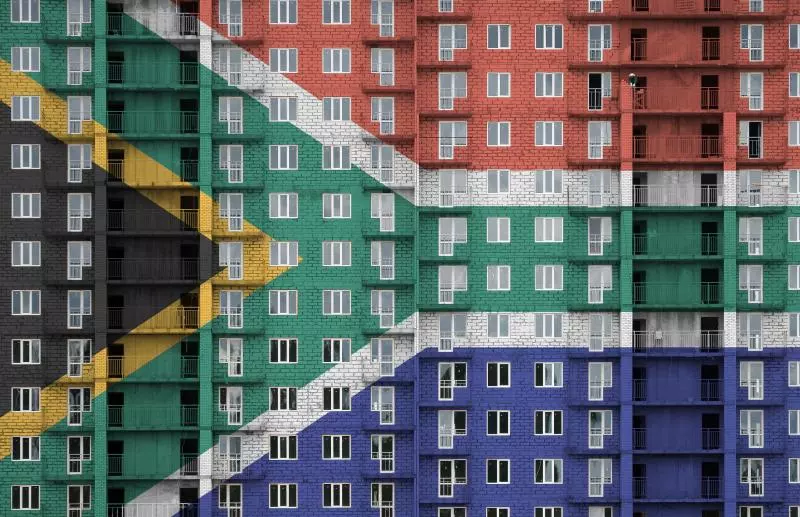 A multi-story apartment building with the South African flag superimposed over it. The building's facade is covered in windows and balconies, and the flag's colors (black, yellow, green, white, red, and blue) are clearly visible.