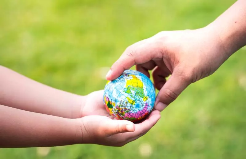 Two hands holding a small globe, with one hand appearing to pass it to the other. The background is a blurred green, suggesting an outdoor setting and symbolising nature.