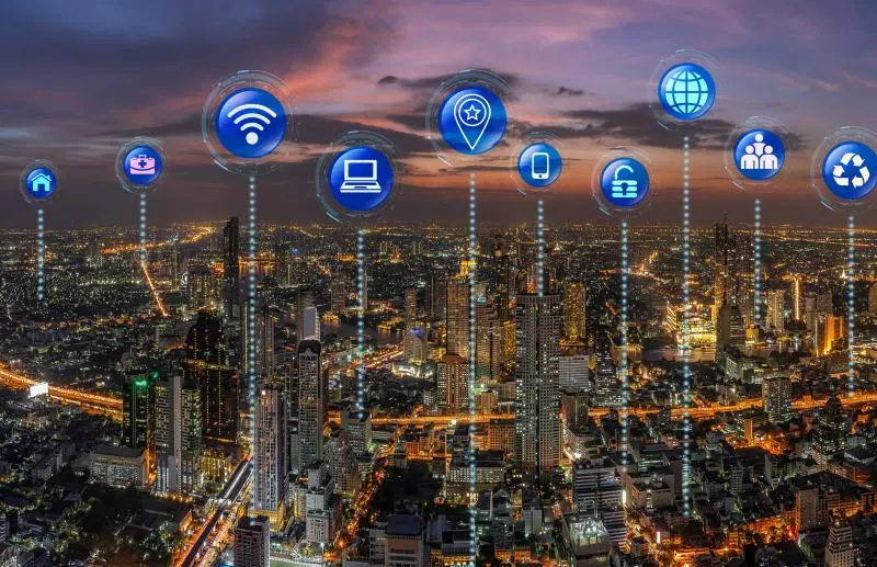 A cityscape at dusk with various technology and lifestyle icons superimposed above the buildings, connected by dotted lines. Icons include symbols for travel, housing, surveillance, internet, computing, location services, mobile communication, education, global connectivity, social interaction, and sustainability.