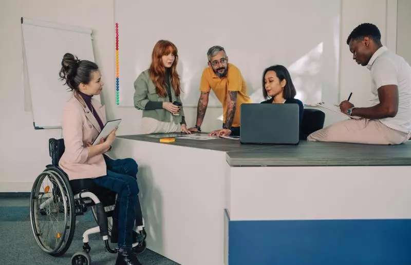 A group of five people collaborating in an office setting. One person in a wheelchair holds a tablet, another stands with a cup, two lean over a table reviewing documents or a laptop, and one sits on the table writing in a notebook. A whiteboard with colorful markers is visible in the background.