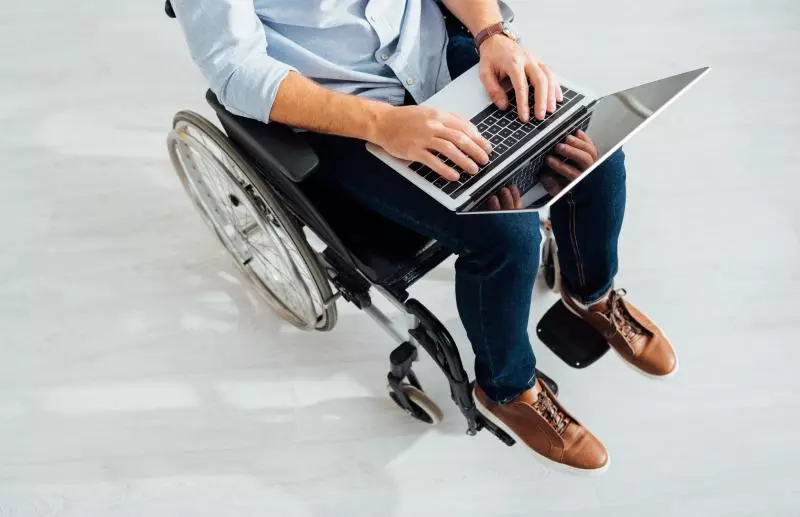 Overhead close-up of a person seated in a wheelchair typing on a laptop; the image shows their hands on the keyboard, legs in jeans, and brown shoes against a light floor.