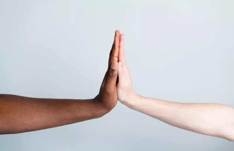 Close-up photo of two outstretched arms, one with dark skin and one with light skin, palms touching in a high-five against a pale gray background.