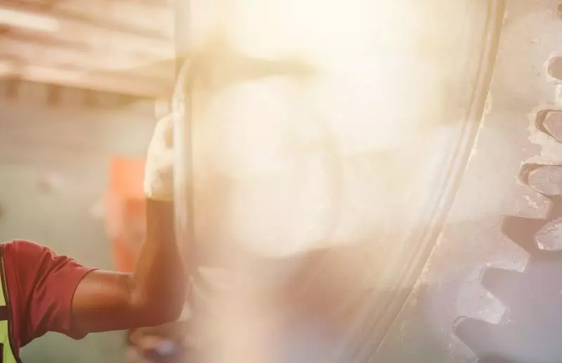 A male factory worker wearing a yellow hard hat, yellow hearing protection around his neck, and a reflective safety vest reaches toward large metal gears inside an industrial workshop; warm sunlight flares in the background.