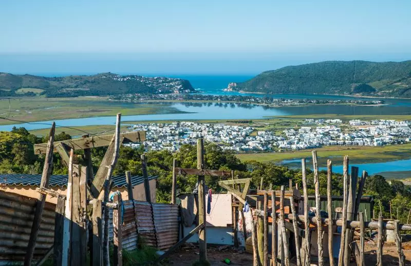 Informal hillside settlement with wooden posts and corrugated metal fences in the foreground overlooking a coastal lagoon and a white seaside town with hills and the ocean in the distance.