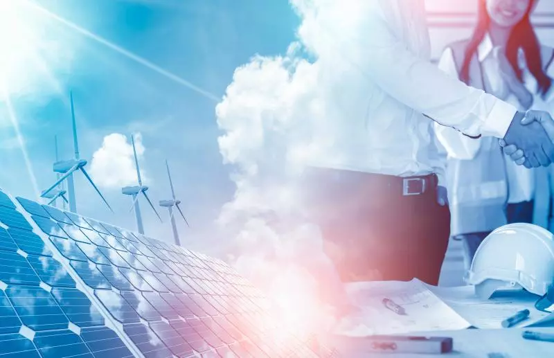 A composite image showing solar panels in the foreground with wind turbines in the distance under a bright sky; at right, businesspeople in shirts shake hands across a table strewn with hard hats, blueprints and a tablet, suggesting a renewable energy project meeting.