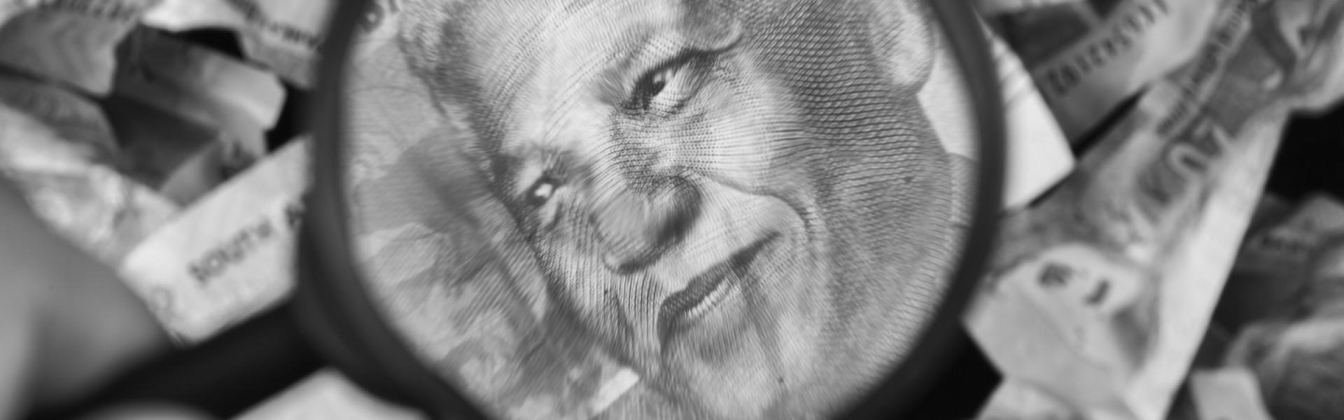 elson Mandela’s face magnified with a magnifying glass on a South African 100 Rand note with crumpled Rand notes in the background